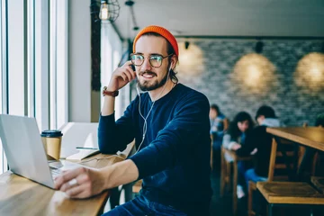 Fotobehang Muziekwinkel Positive male meloman in optical spectacles feeling good from received friendly audio message, happy hipster guy sitting in coffee shop with laptop and listening music playlist via earphones