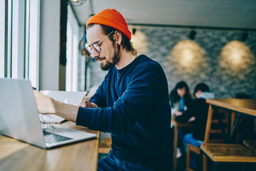 Millennial hipster guy in trendy spectacles sitting at cafeteria table with modern laptop computer,...