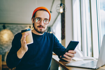 Amazed hipster guy with shocked expression on face checking mails via smartphone device during coffee break, surprised man connected to 4g wireless on cellphone for chatting in social networks