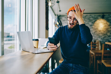 Wonder hipster guy in optical eyewear feeling shocked from received text message about mistake in...