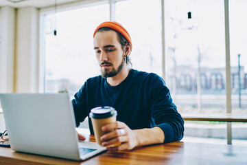 Tired caucasian male student drinking coffee completing coursework on laptop computer in coworking space, concentrated hipster guy working online on project drink caffeine finish before deadline.