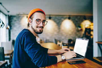 Portrait of happy male freelancer in optical eyewear for vision correction smiling at camera during break from web working online, cheerful hipster blogger sitting in cafe with mockup laptop computer - obrazy, fototapety, plakaty
