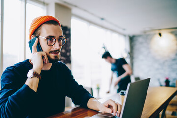 Caucasian hipster guy in trendy bennie making telephone call to customer support talking about laptop computer, man having smartphone conversation checking banking balance for making transaction. - obrazy, fototapety, plakaty