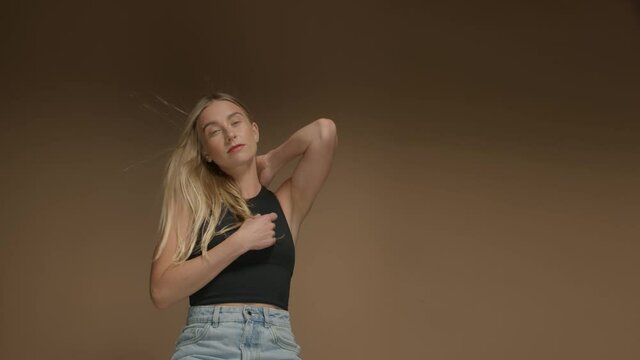 young blonde woman with long hair and blue eyes in studio on beige background poses to the camera touches hair lift hands up