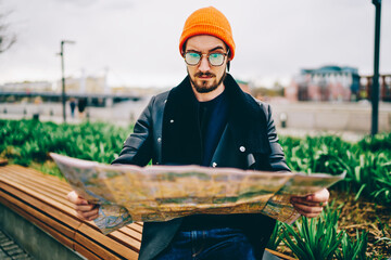 Confused male tourist in spectacles and orange hat looking for route on map can't understand...