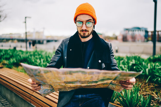 Confused male tourist in spectacles and orange hat looking for route on map can't understand location, shocked hipster guy checking destination stressed with get lost in city during travelling.
