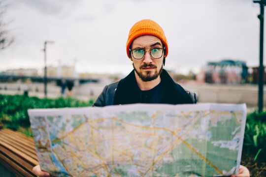 Portrait of puzzled tourist 20s holding paper location map confused with lost direction and destination, Caucasian hipster guy in trendy spectacles looking at camera during city sightseeing