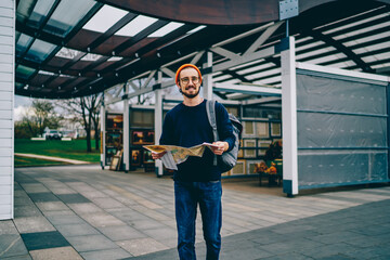 Portrait of happy Caucasian male tourist in stylish glasses smiling at camera during travel sightseeing, good looking backpacker holding paper map for search location direction and walking in city