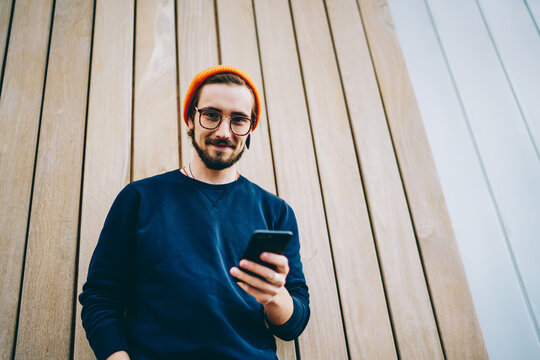 Half length portrait of millennial user in optical spectacles looking at camera while posing at urban publicity area,handsome hipster guy holding modern smartphone technology connecting to 4g internet