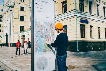 Young male traveler using modern smart board with map and useful information for tourists, caucasian hipster guy searching train on electronic device on street with wifi connection and touchscreen .