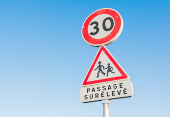 Round 30 kilometers per hour speed limit road sign in a children school area. Written in french "passage surélevé" (speedbump) Caution. Blue sky in the background.