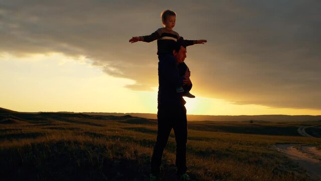Silhouette of a happy family at sunset. The son sits on the shoulders of his father and depicts the flight of an airplane. The family is relaxing in the fresh air, dreaming of traveling.