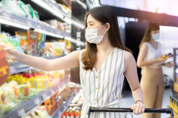 Asian woman wearing face mask push shopping cart in suppermarket . Girl choosing, looking somethings to buy at shelf during coronavirus crisis or covid19 .happy lifestyle or shopaholic concept.