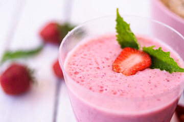 Cocktail with strawberries and milk in a glass decorated with mint and strawberries. Nearby is a pink cup with Hercules flakes, and loose strawberries on a white wooden background. 
