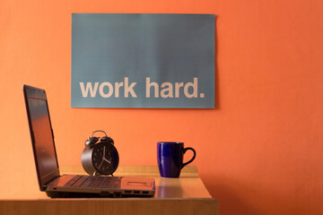 A home workspace including a laptop kept on a desk along with an alarm clock, a coffee mug, and a work hard poster on a wall.