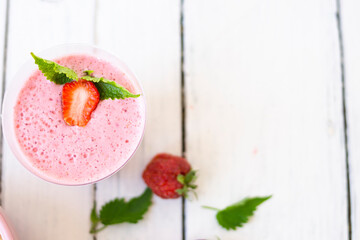 Cocktail with strawberries and milk in a glass decorated with mint and strawberries. Nearby is a pink cup with Hercules flakes, and loose strawberries on a white wooden background. 