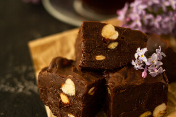 A piece of homemade chocolate cake on crumpled craft paper, with nuts on a black texture table. A cup of tea and lilac flowers decorate a delicious tea party.