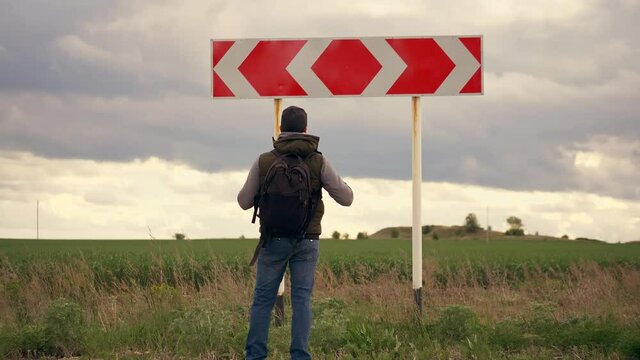 Choice of a way. Man tourist with a backpack comes up to the road direction sign and chooses the path. The problem of choice, decision making.