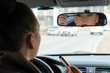 Beautiful female driver makes-up in the car and putting mascara on her eyes. Young lady looking at rearview mirror in the car. Focus on the mirror