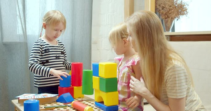 Young mother and two cute daughters playing with colorful plastic blocks building a tower together at home. Games and preschool education.