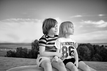 Two boys sitting on hay bale in the meadow, hugging, smiling and watching sunset. Holidays in the countryside. Black and white photo.