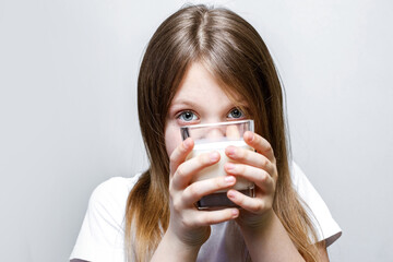 The girl looks out mischievously from behind a glass of milk. Close-up of a pretty girl holding a white milk in a transparent glass.