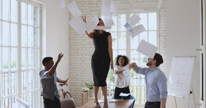 Excited diverse business team and happy female leader having fun throwing tossing papers up in office room on friday, euphoric woman boss winner dancing on table celebrate success at corporate party