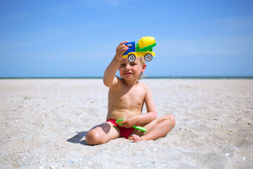 Little charming boy sit at sea beach sand, hold plastic scoop and show concrete mixer toy plastic...