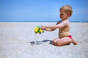 Little kid boy play at sea beach, pour out sand from toy plastic concrete mixer car, wearing red...