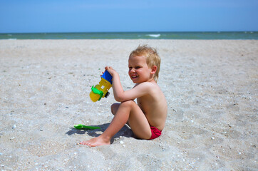 Little happy tanned child boy 3-4 years old sit at beach sand and play with plastic toy car and scoop with sea view, family vacation
