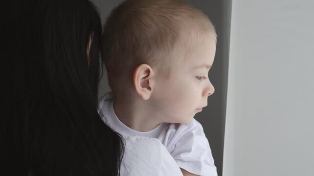 A dark-haired mother holds her son in her arms while standing at the window. The view from the back. The boy looks out the window.