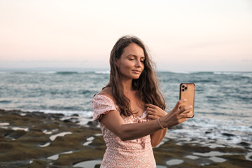 Fototapeta na wymiar Beautiful girl preens looking at the phone on the beach on a background of the sea. A nice young girl straightens her hair while looking in a cell phone.