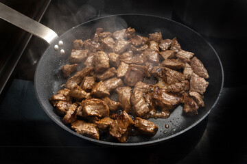 Steaming beef cubes are roasted in a pan on a black stove for stew or goulash, cooking at home concept, selected focus