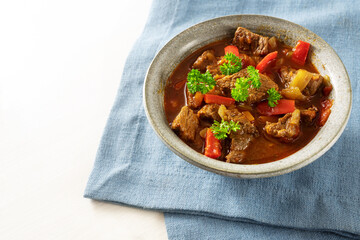 Goulash or stew from beef, onions and red bell pepper with parsley garnish in a bowl on a blue napkin and a white table, copy space