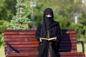 Arab woman in black national dress is reading a book in city park. A migrant from the Middle East...