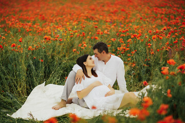 Happy couple - pregnant woman and her husband lying on white plaid in red poppy field. Future...