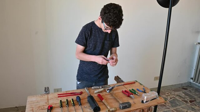 A Caucasian boy is intent on ordering work tools on a table: screwdrivers, hammers, pliers, utility knife and protective glasses can be seen.