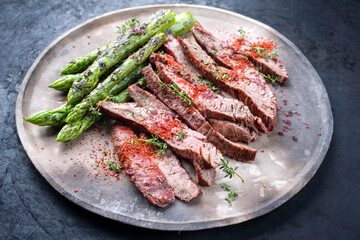 Traditional barbecue sliced dry aged wagyu flank steak with green asparagus and chili powder offered as close-up on a rustic modern design plate