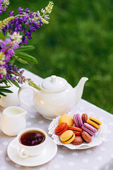 A cozy romantic breakfast in the fresh air. A vase with lupine flowers, a teapot and a cup of tea, a cake with macaroons on the table. Congratulations on Mother's Day or Women's. Soft selective focus.