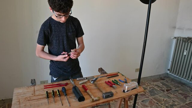 A Caucasian boy is intent on ordering work tools on a table: screwdrivers, hammers, pliers, utility knife and protective glasses can be seen.