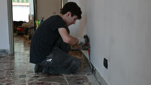 Masonry work in the apartment: a Caucasian boy is wearing work gloves and is using hammer and chisel to demolish the old baseboard. 