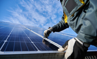 Close up of man technician in work gloves installing stand-alone photovoltaic solar panel system under beautiful blue sky with clouds. Concept of alternative energy and power sustainable resources. - obrazy, fototapety, plakaty