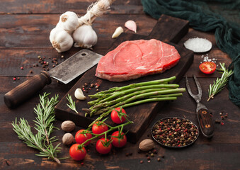 Slice of fresh raw barbeque braising beef steak on chopping board with asparagus and garlic with tomatoes and salt with pepper on wooden background.