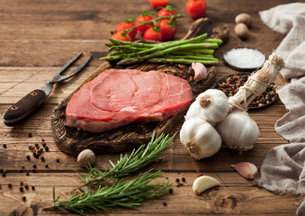 Raw slice of beef braising steak on chopping board with asparagus, rosemary and garlic with tomatoes and salt with pepper.Perfect meat for barbeque on wood.
