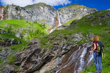 Lady hiker looking at waterfall on mountain. One person with backpack outdoor activity in scenic alpine landscape, summer vacation on the Alps, fitness wellbeing freedom