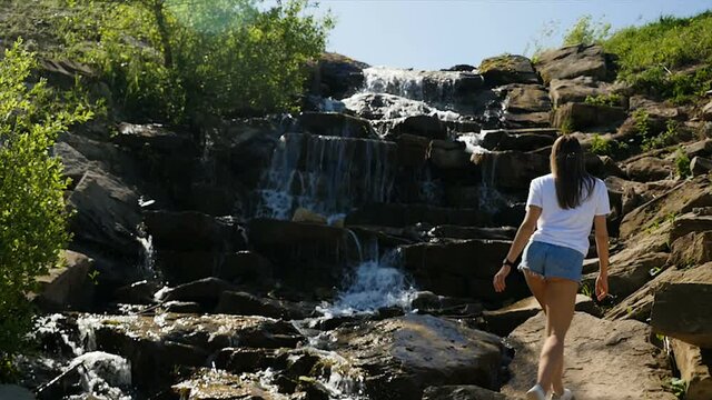Young lady climbing rocks near waterfall. Young slim woman in t shirt and shorts climbing wet stones then stopping and resting near waterfall with clean water on sunny day