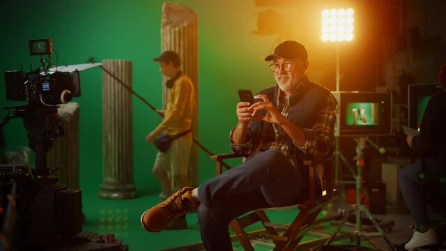 Prominent Successful Director Sitting in a Chair on a Break Using Smartphone. On the Studio Film Set with High-End Equipment Professional Crew Shooting High Budget Movie. Zoom In Looking at Camera