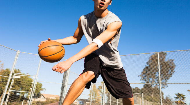 Male teen basketball player dribbling the ball on an outdoor court during a sunny day. Man athlete playing streetball.