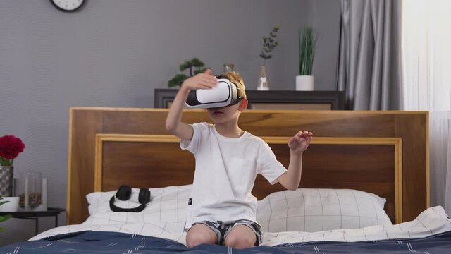 Handsome satisfied small boy in virtual reality headset sitting on bed and playing game on imaginary screen