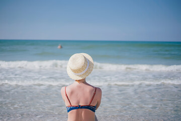 Fototapeta na wymiar girl in a swimsuit and a straw hat sits on the sand by the sea 